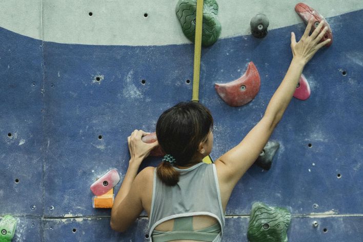 Back view of unrecognizable slim female mountaineer in sportswear with harness climbing artificial rock with grips